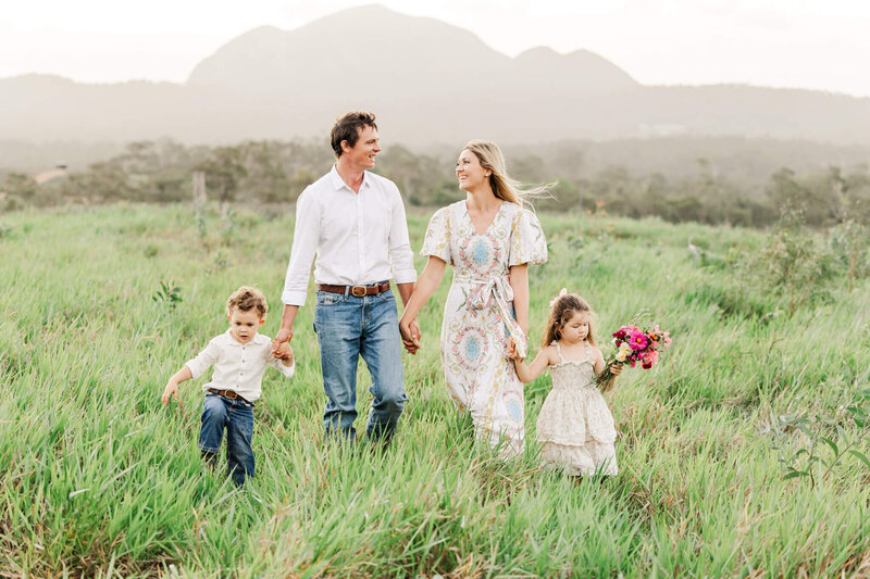 Stylishly dressed family of four walking through green grass with a mountain range as a backdrop. Dad and Mum holding hands of their son and daughter while they glance happily at each other.