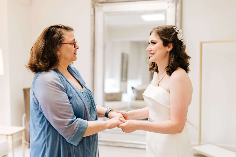 A bride in a white dress and a woman in a blue outfit hold hands and smile warmly at each other in a bright room with a large mirror in the background.