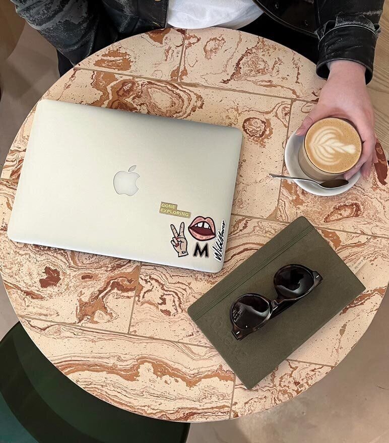 Martina Menzini working on laptop at marble table in cafe, with coffee, sunglasses, and planner