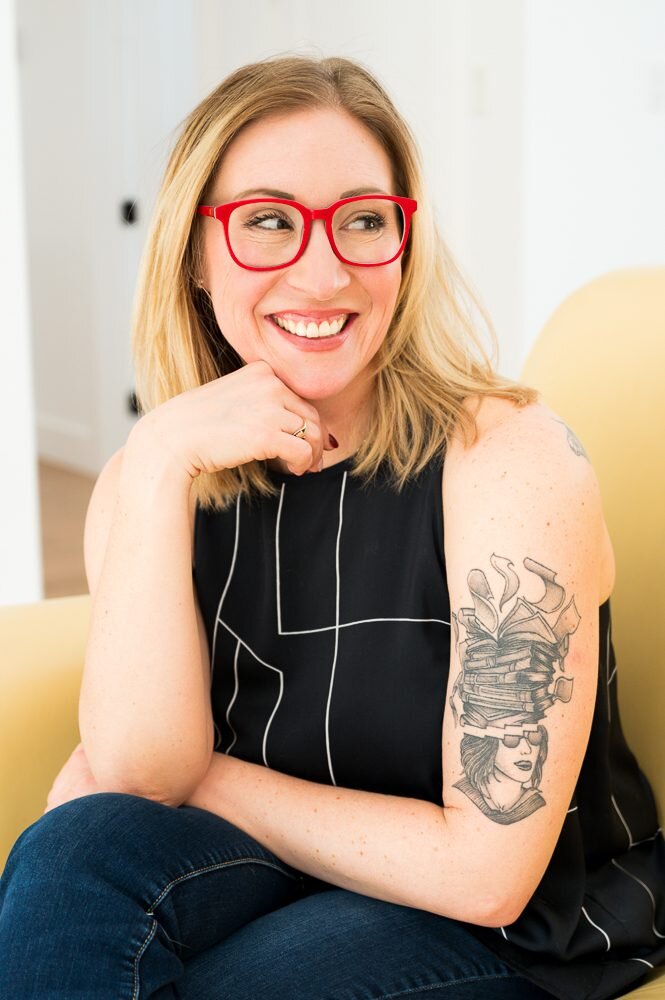 A woman with glasses and tattoos sits comfortably on a chair, exuding a relaxed and confident demeanor.
