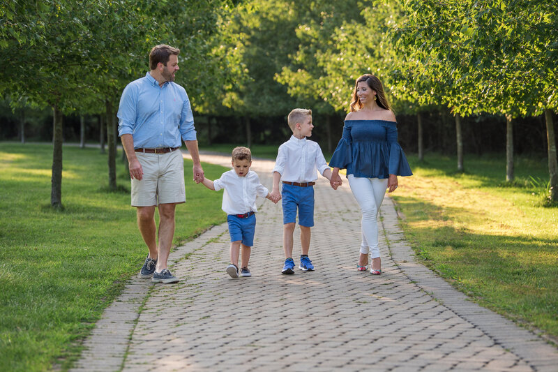 Family of 4 walking together and holding hands on a stone pathway at a park.