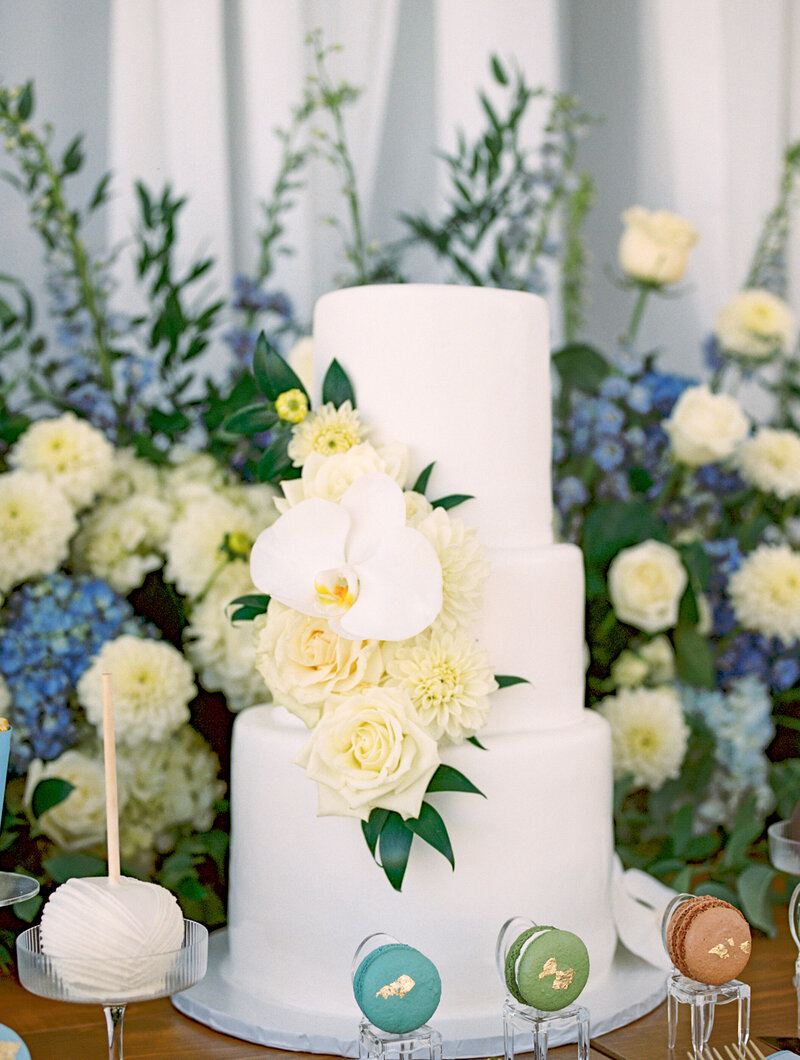 White three-tier wedding cake with ivory roses and orchid flowers, surrounded by blue and white florals | Cleveland wedding photographer detail photo