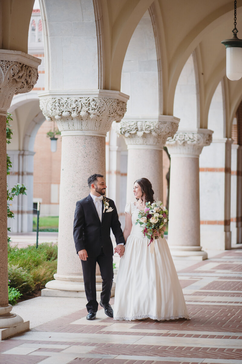 Bride in a strapless ballgown holds a bouquet while walking hand in hand with the groom in a tuxedo along a sunlit courtyard during their wedding.