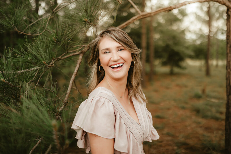 Allyson Blankenburg, Houston wedding photographer, poses for a headshot wearing an earthy toned dress, among pine trees.