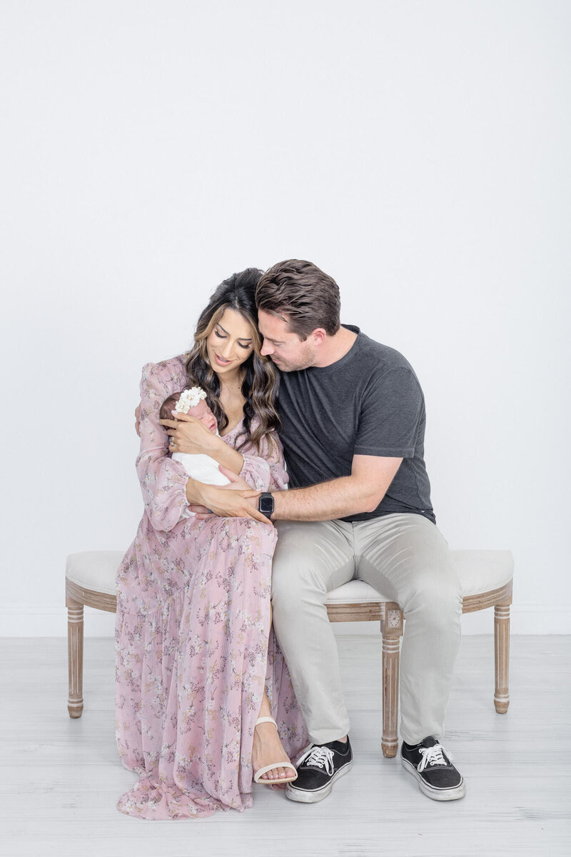 A couple sits close together on a bench, lovingly cradling a newborn baby wrapped in a white blanket. The woman wears a pink floral dress, and the man is in a black shirt and gray pants. The background is a plain white wall.