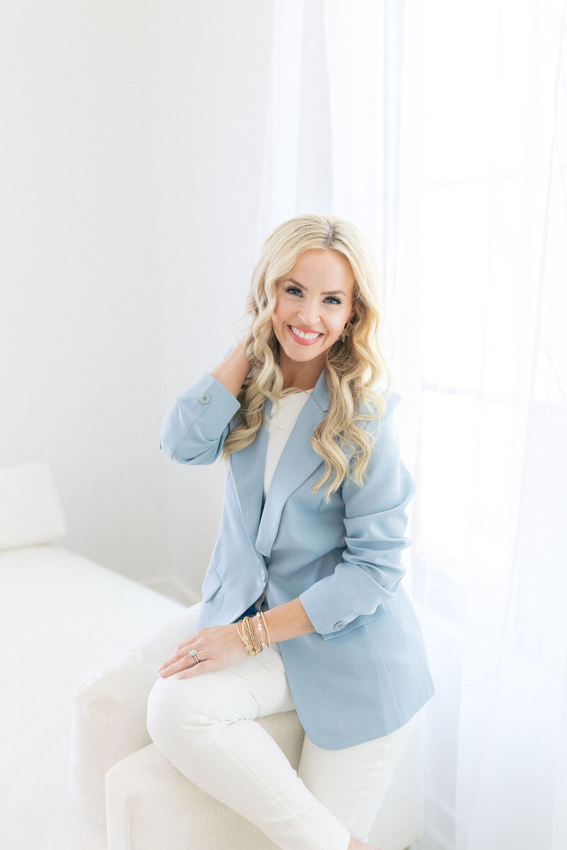 Woman in blue tailored jacket sits on white couch and smiles