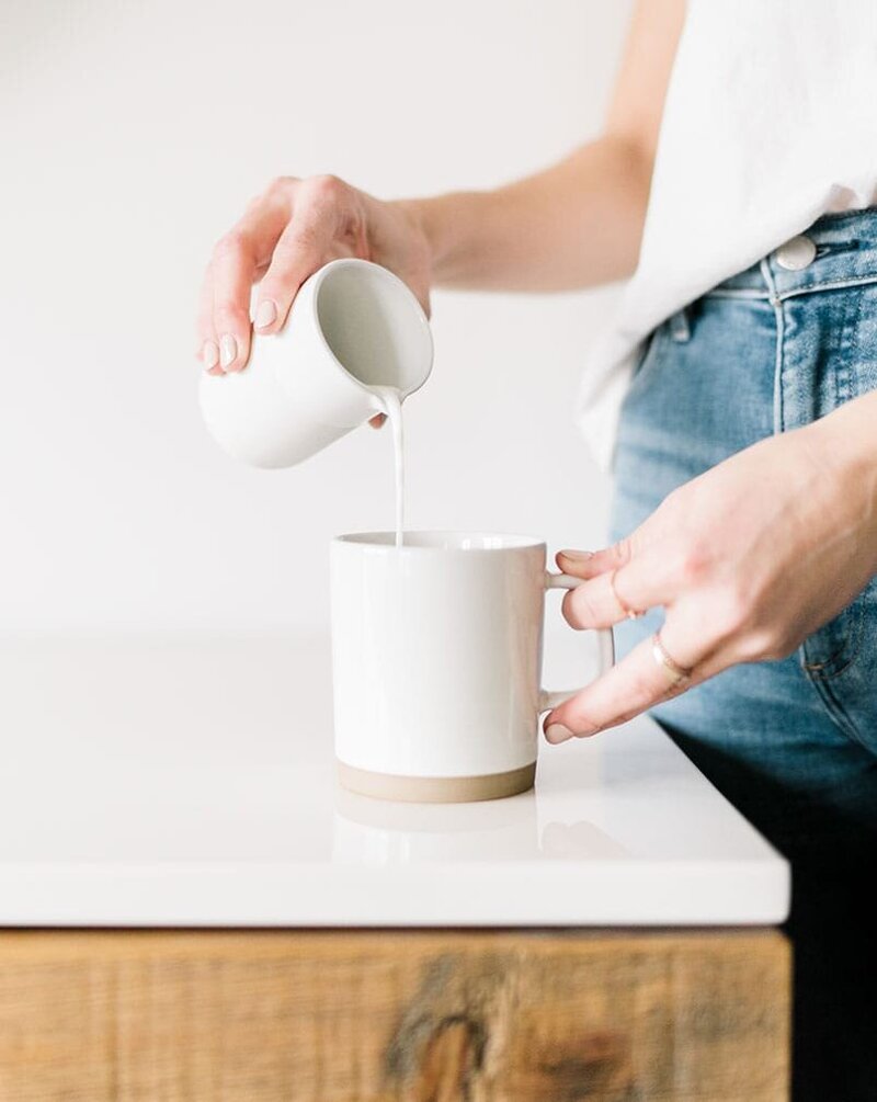 Close-up of a woman pouring cream into a coffee mug at a white kitchen counter, representing accuracy in Xanthe Bookkeeping’s business values.