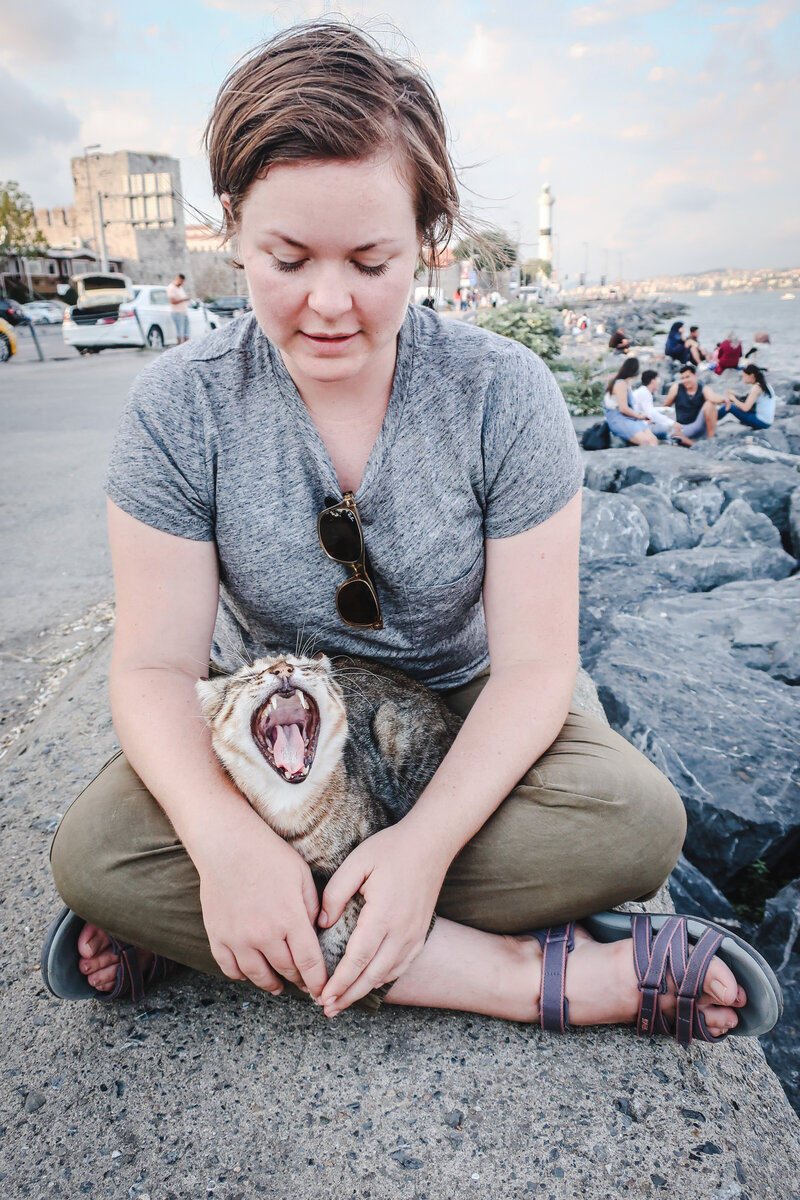 Michelle-Marie Gilkeson sits by the sea in Turkey  looking down at a yawning cat in her lap