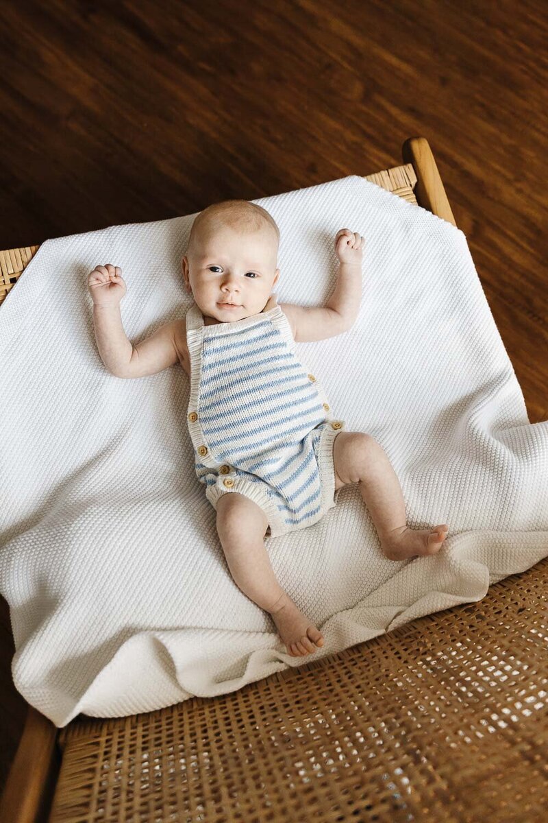 A baby lies on a white blanket, wearing a striped romper, with arms slightly raised on a wooden woven chair during their toronto newborn portrait session. 