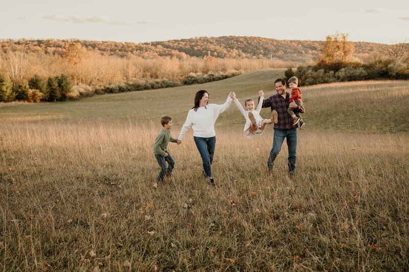 Family swinging young daughter in a fall field with tall grass and autumn mountains in the background
