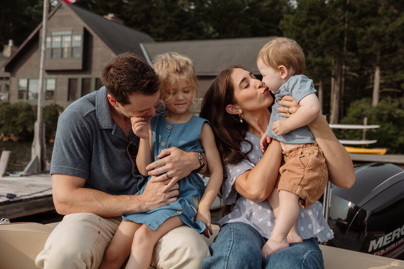 A family of four snuggles up on a boat with their family cabin in the background. A mother and father hold their children close — the mother kisses their baby boy as he stands on her lap, while the father holds their daughter in his arms. Dressed in neutral colors and lake-weather layers, they glow in the golden light. Captured with the finesse of a New England photographer, the image radiates fine art elegance and a tender, documentary-style warmth.