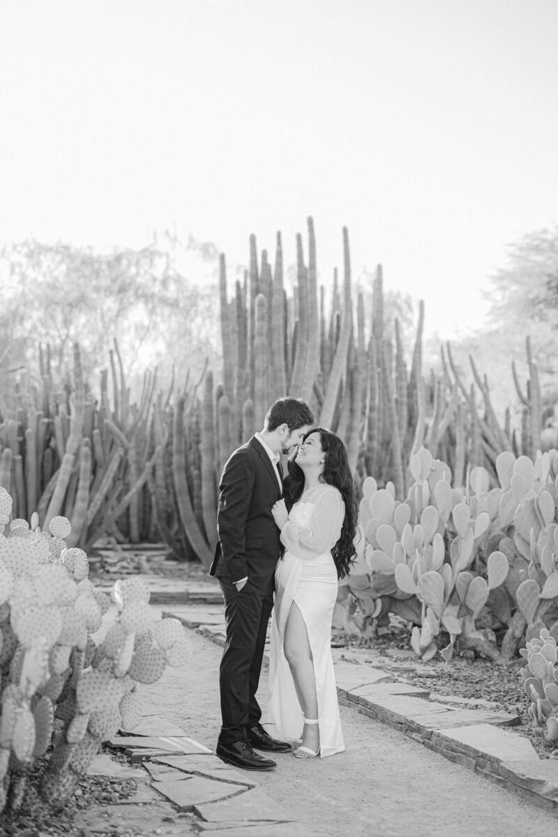 A couple, dressed in classic attire, embraces amidst a botanical garden, gazing at each other with eyes full of love.