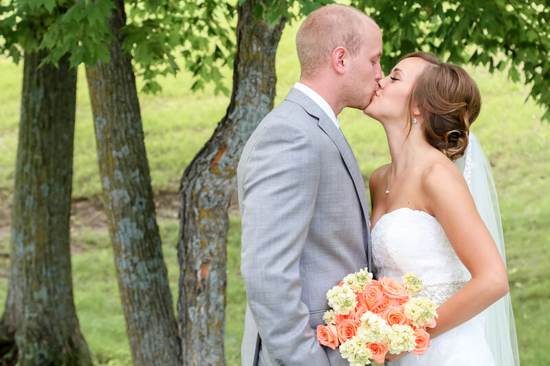 A white couple wearing wedding attire is kissing under a tree