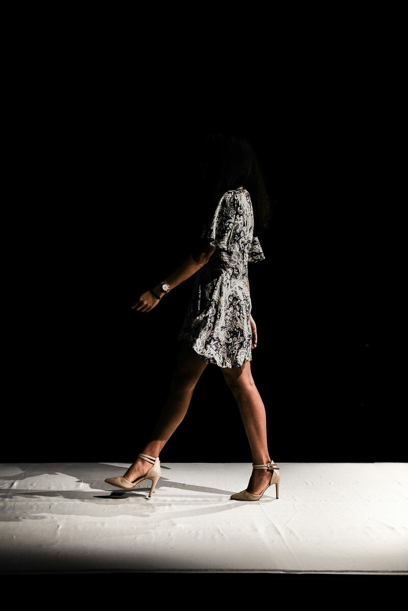 Model in a dress and heels walking on a white runway in front of a dark black background
