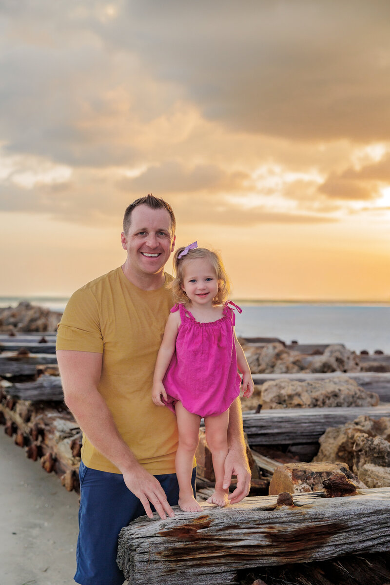 Father and daughter posing on  a jetty on the beach with the sunset behind them.