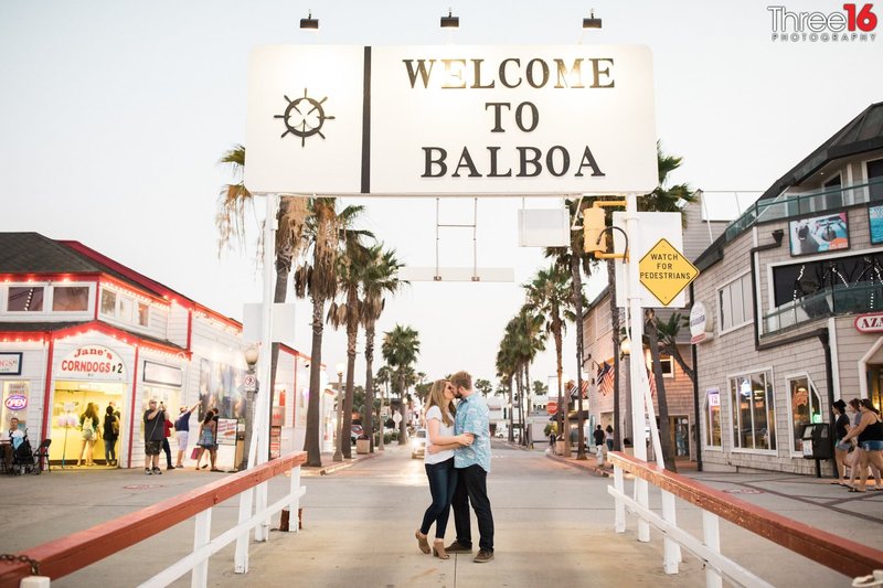 Engaged couple kiss under the Welcome to Balboa sign on Balboa Island in Newport Beach