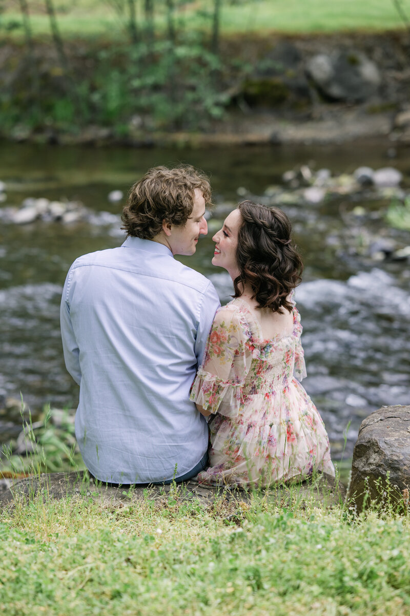 man and woman sit facing river for engagement
