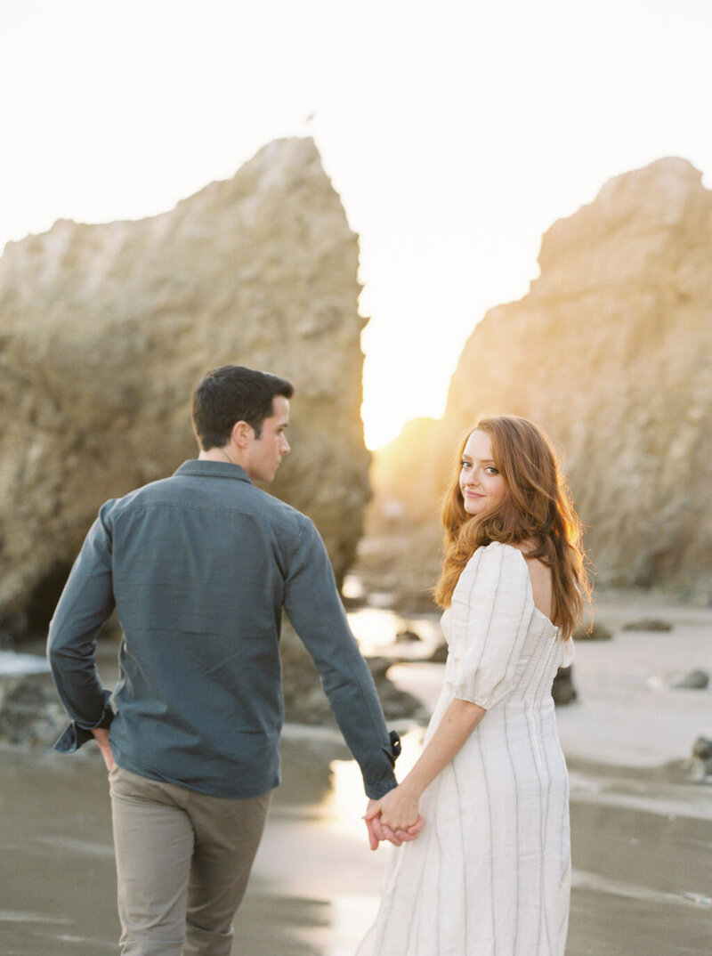 engaged couple holding hands walking on beach next to cliffs