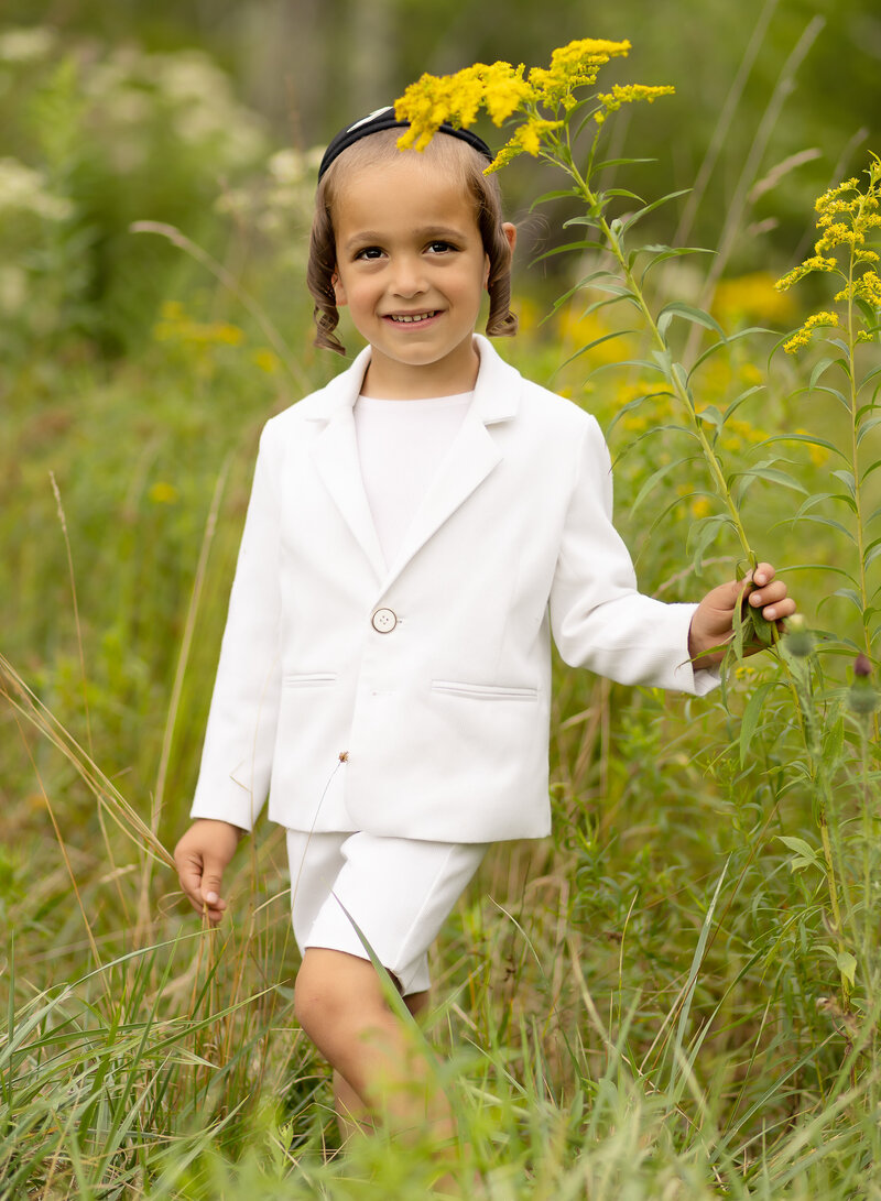 Little boy in a white suit jacket and shorts is standing in a field for an outdoor family photoshoot. He is smiling at the camera. Captured by best Brooklyn, NY family photographer Chaya Bornstein.