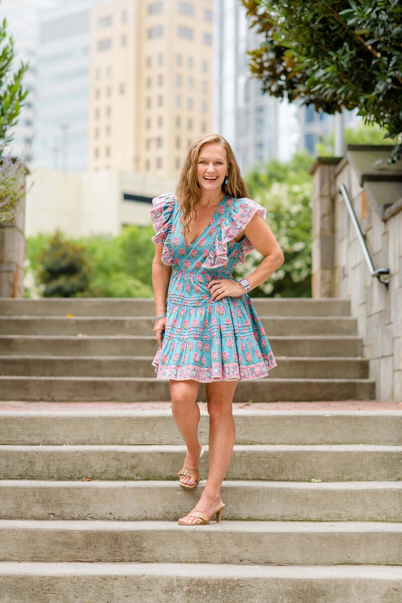 photographer dressed in teal and pink standing on stairs in uptown charlotte with her hand on her hip laughing