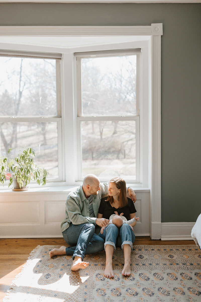 Mother and father sitting with newborn baby in front of sunny window on the floor