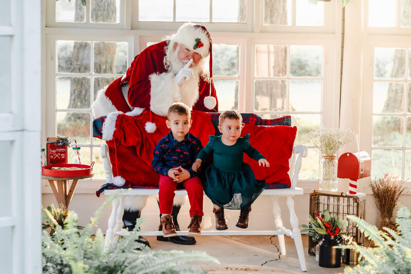 2 toddlers sit together on a bench in a sun porch. Santa is behind them with his finger to his lips.