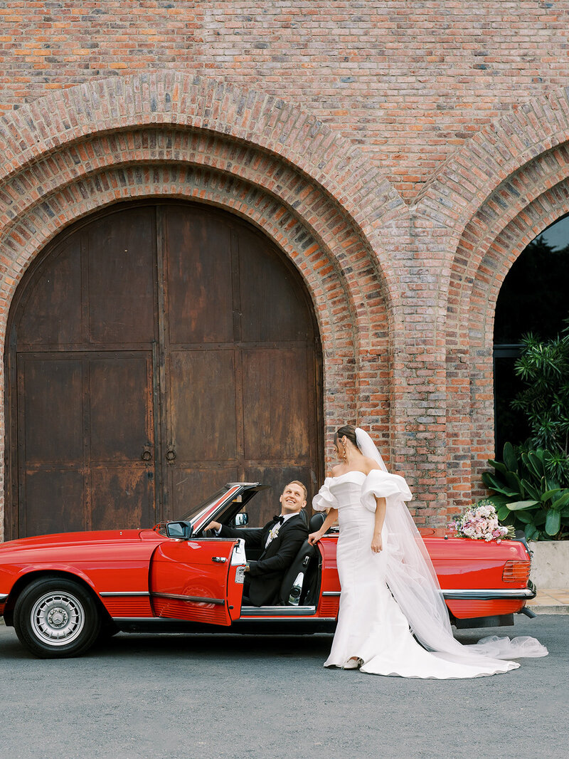 Bride and groom in the getaway car. This editorial shot has the bride standing outside of the car and the groom at the driver's seat looking up at her.