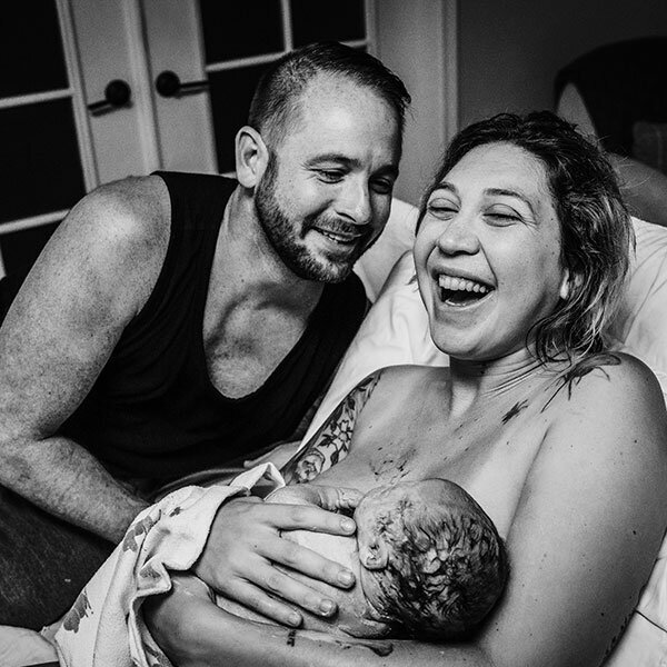 mother and father laugh joyously in bed immediately after the birth of their baby