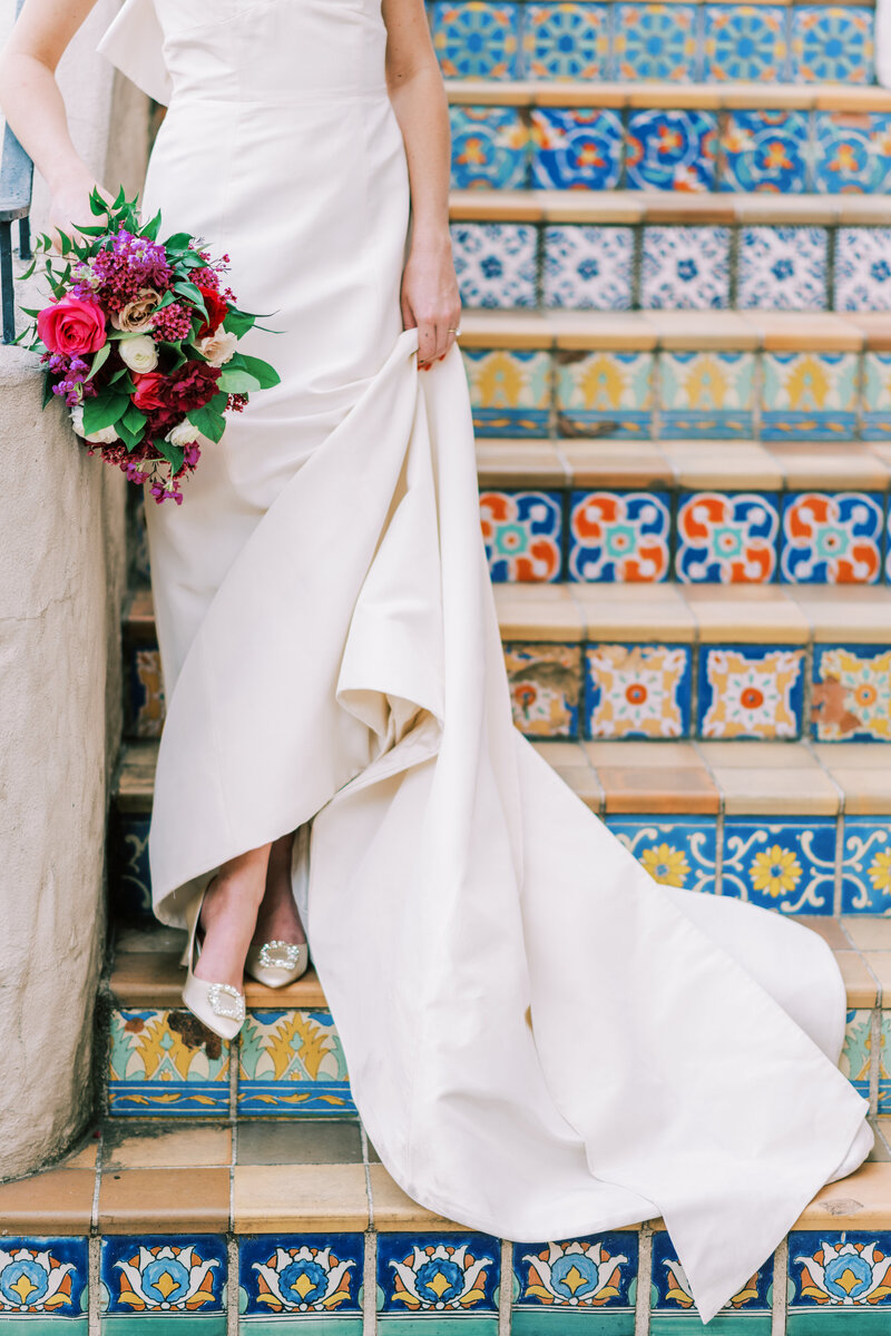 Close up image of the bottom of a bride's dress while she stands on colorful patterned stairs