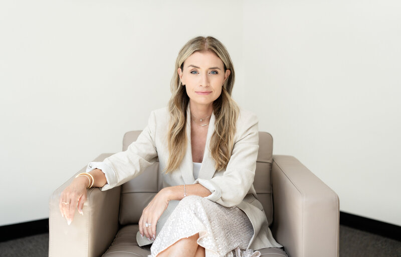 Corporate coach Wendy Tamis Robbins wearing a white blazer sitting on a grey armchair