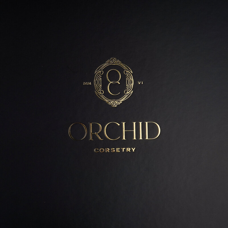 Gold embossed Orchid Corsetry logo shown on a black textured surface