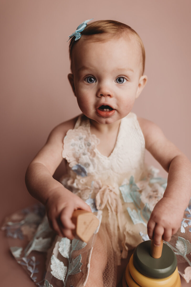 Baby girl sitting against a pink background playing with a wooden toy.