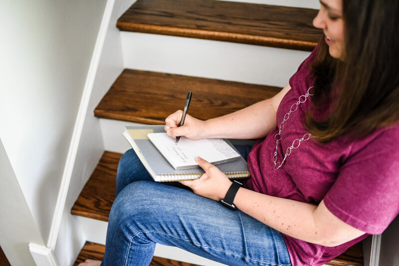 Danielle Hardesty sitting on hardwood stairs smiling and writing in notebook
