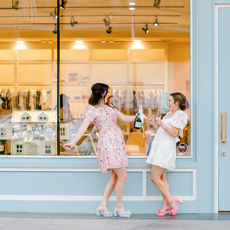 Two women in dresses smiling and popping champagne outside of a store