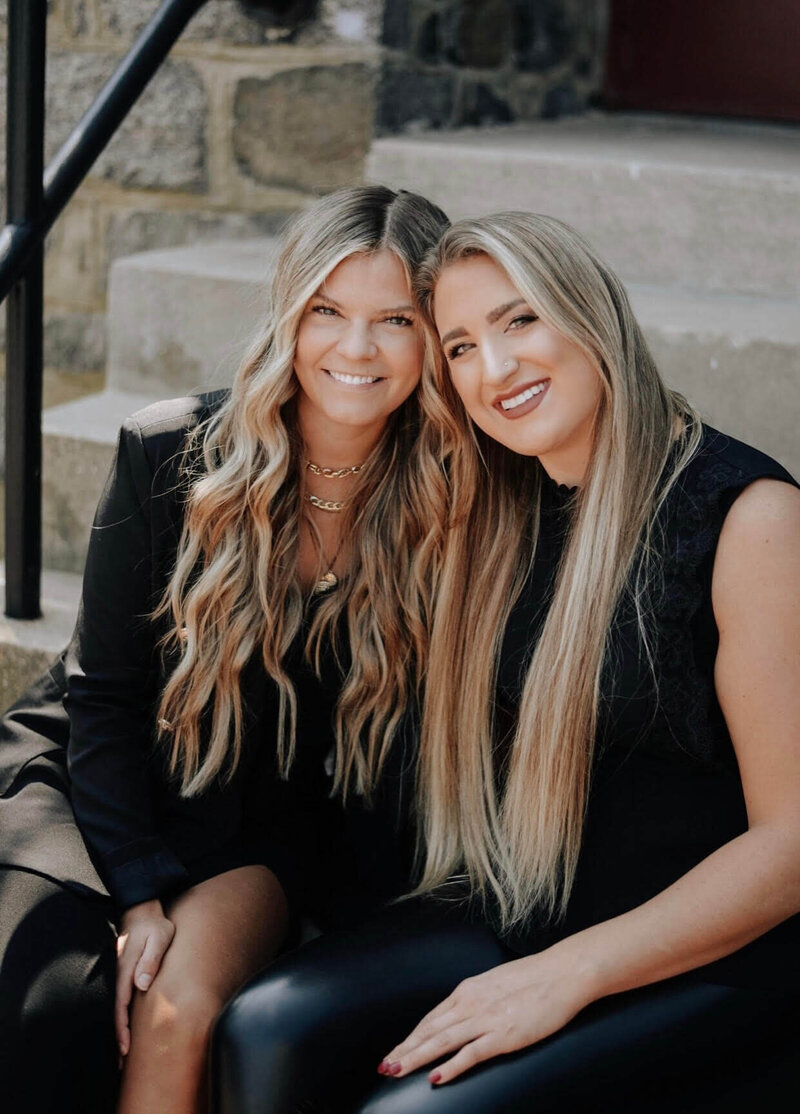 Two blonde female hair extensions stylists smiling with heads close together wearing black