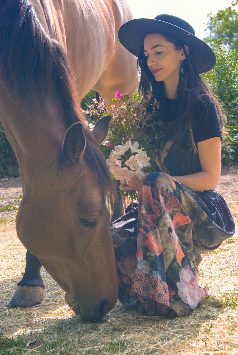 Animal Communicator kneeling beside grazing horse holding flowers. 