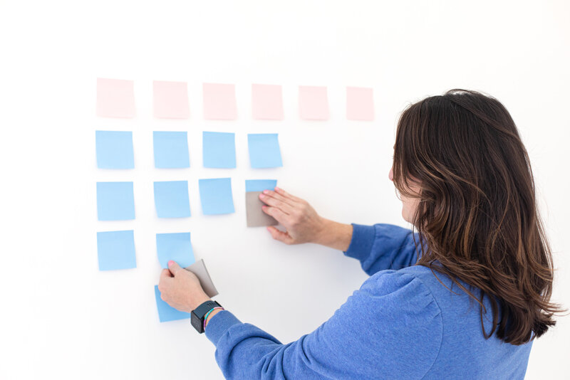 social media strategist and course creator dressed in a blue jacket, she is sticking light pink and blue post it's to the white wall. in her left hand she is holding a black smart watch