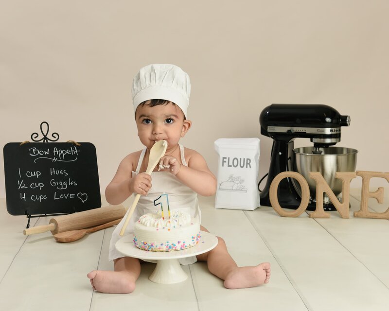 A baby boy dressed as a chef with a white apron and hat, sitting next to a cake and surrounded by baking tools and a "ONE" sign in a cooking-themed cake smash photoshoot.