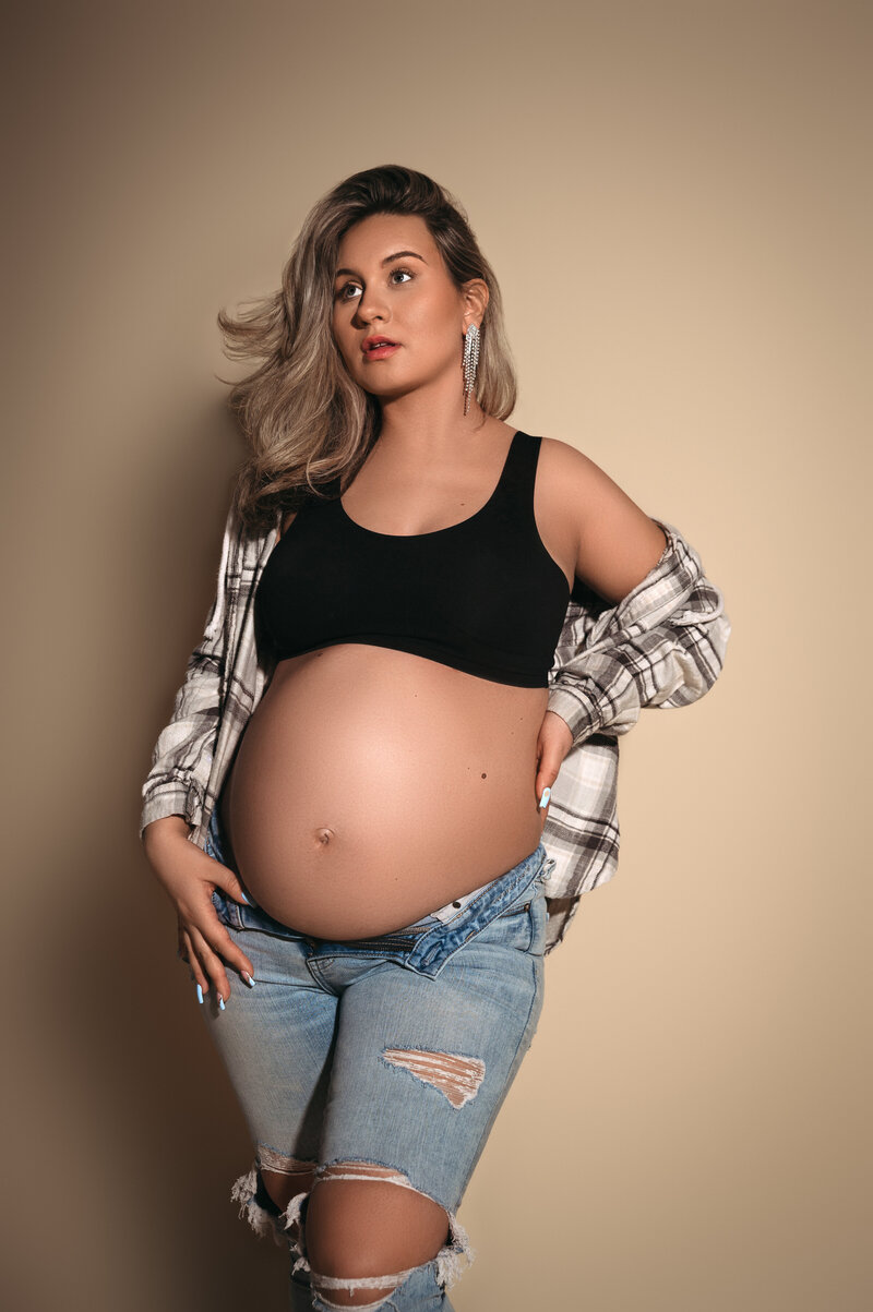 Beautiful pregnant woman holding her baby bump wearing ripped jeans and plaid open button down shirt  and looking off into the distance