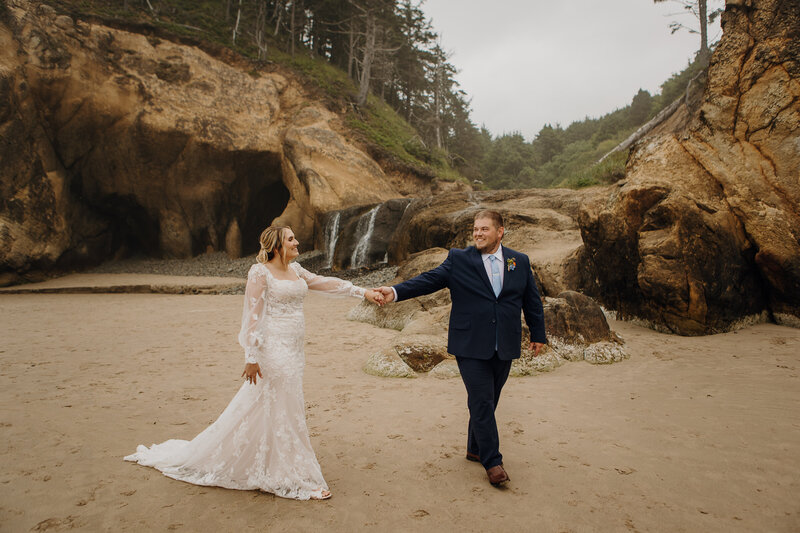 a newly eloped couple in wedding gear walk hand in hand on the sandy shores of Hug Point State Park at Cannon Beach. 