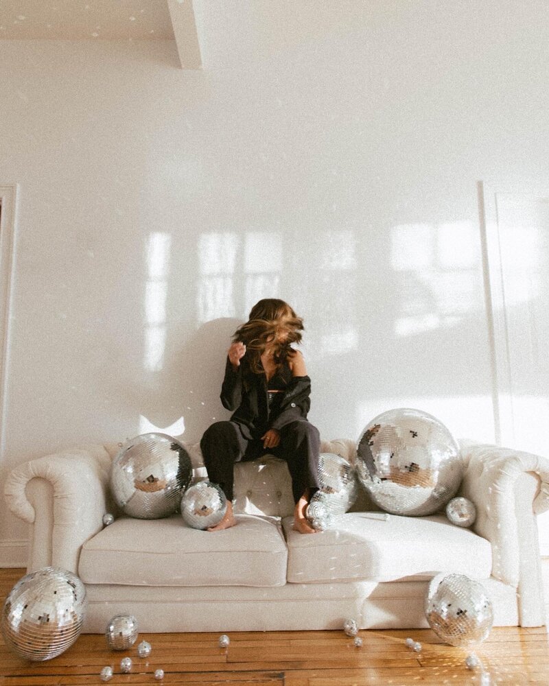 Wedding and portrait photographer, Allie, sitting on a couch in a black power suit surrounded by disco balls
