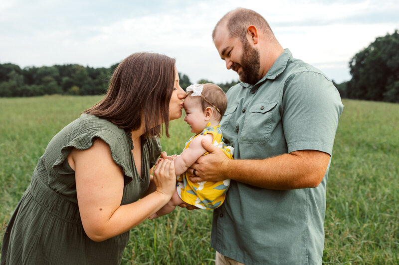 family of three in a field with dad holding baby and mom kissing her forehead photoshoot with jennifer mosier photography