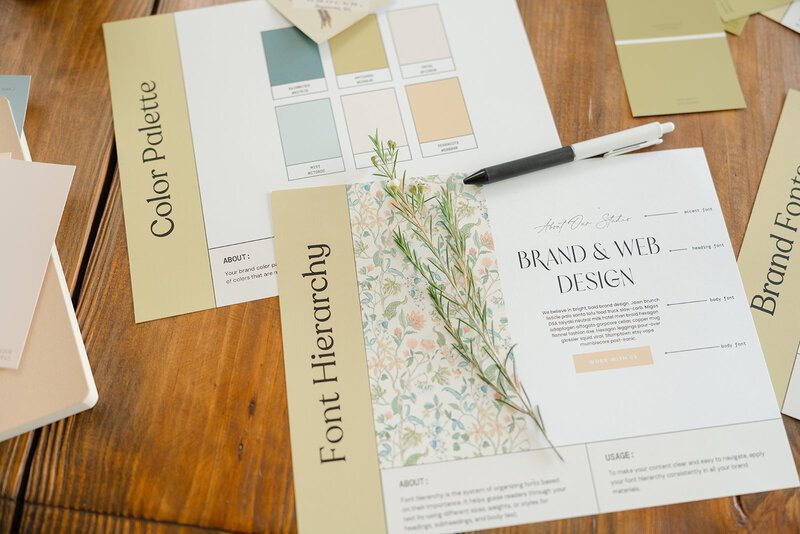 Pages from a brand guidelines book displayed on a wooden table with a piece of green filler floral laying on top