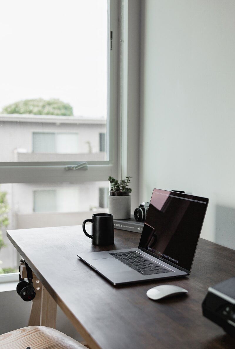 a light-filled home office with a black laptop, coffee mug, mouse,  plant, and camera