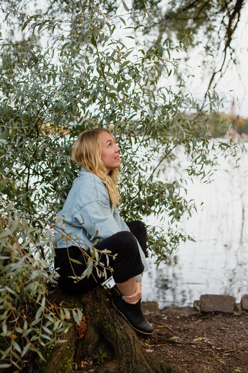 mielen paremminvoinnin ja kehomieliyhteyden valmentaja  istuu puunrungon päällä järven rannalla puunoksien alla ja katsoo eteenpäin