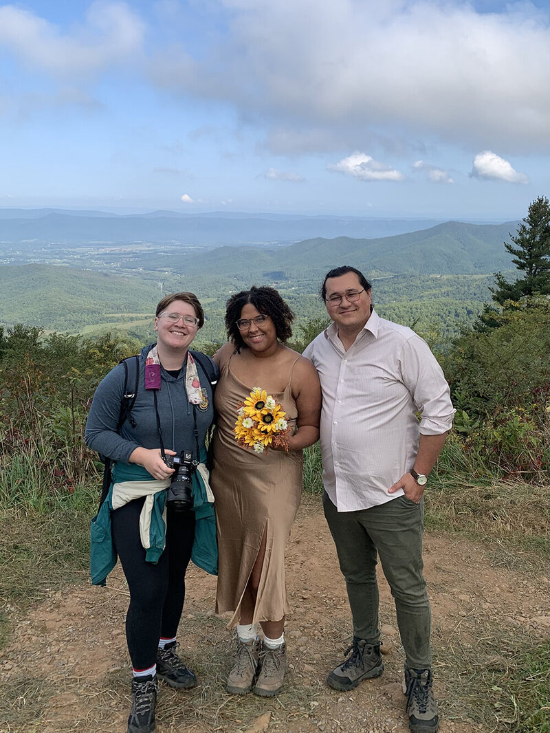 A Virginia elopement photographer stands next to an elopement couple on their wedding day. The trio stand at a popular photo spot at Jewel Hollow Overlook in Shenandoah National Park.