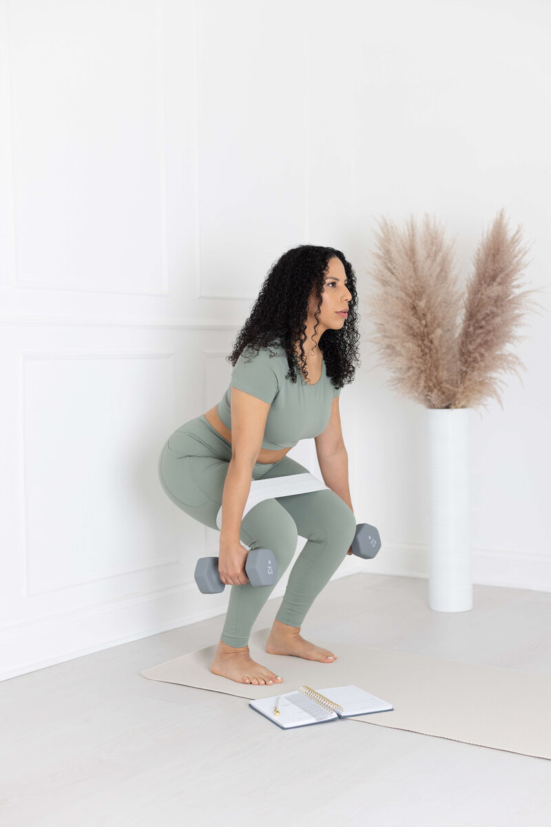 woman in green active wear squatting with gray dumbbells  and fitness journal open on the floor