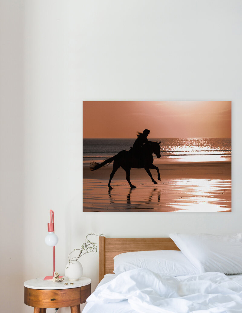 A Gallery Block wall art showing a horse and rider galloping on a beach hanging over a bed