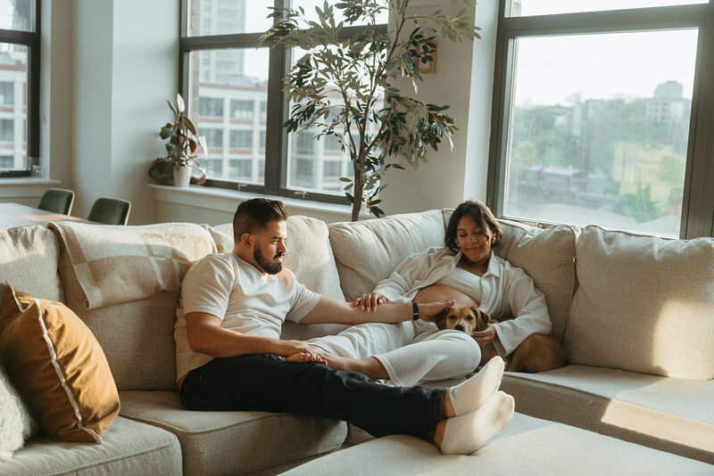 pregnant wife sits on couch with husband and dog during maternity photos captured by NYC maternity photographer Elsie Goodman 