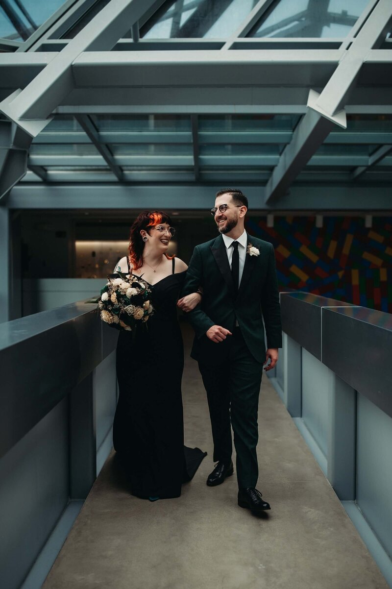 Bride in black dress and Groom in Green suit walk at art museum for moody wedding.